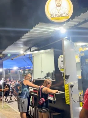 Quiosque do Seu João com o letreiro redondo aceso e clientes no balcão
